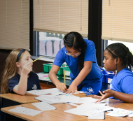 Kids sitting in groups of desks watching and listening to a teacher.  Image by Kenny Eliason on Unsplash.